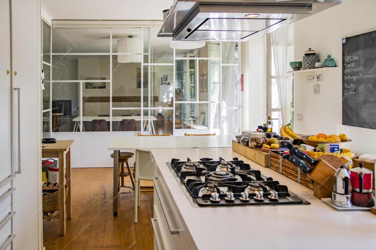 Cucina con fornelli a gas, vetrate industriali dagli infissi bianchi e vista sul salotto