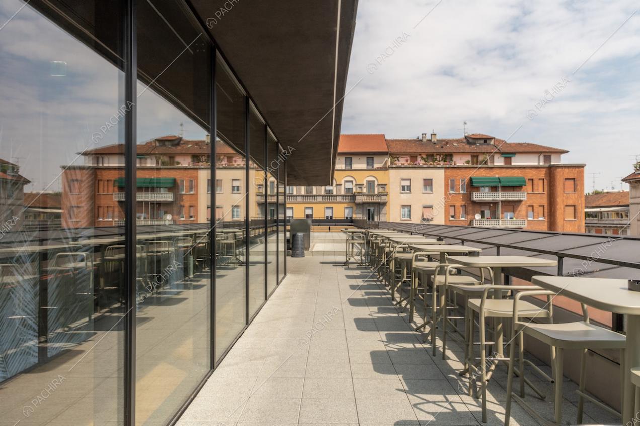 Terrazza con tavolini e sedie in metallo, vetrate con scorcio sugli interni e vista su Milano