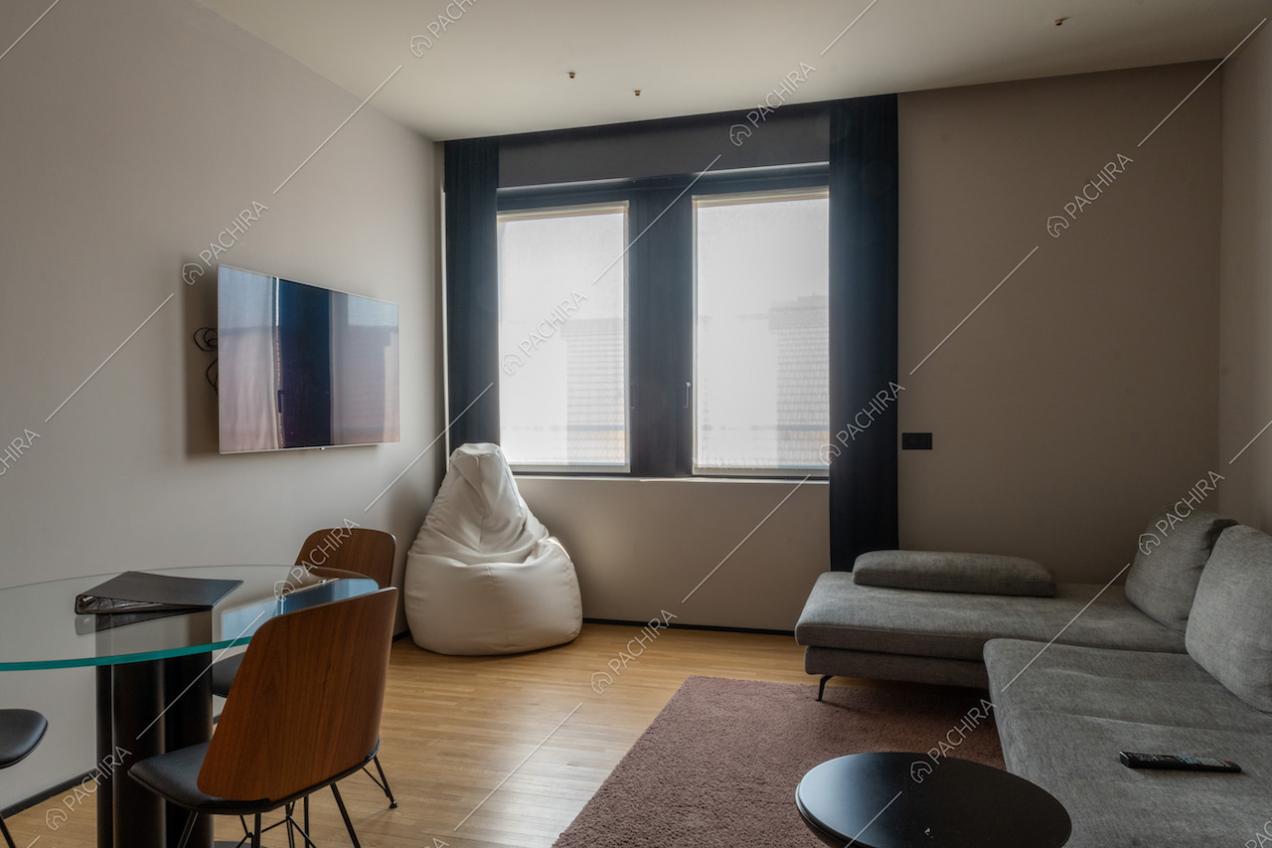Salotto con tavolo dal piano in vetro, pouf bianco, televisore a muro e divano grigio angolare