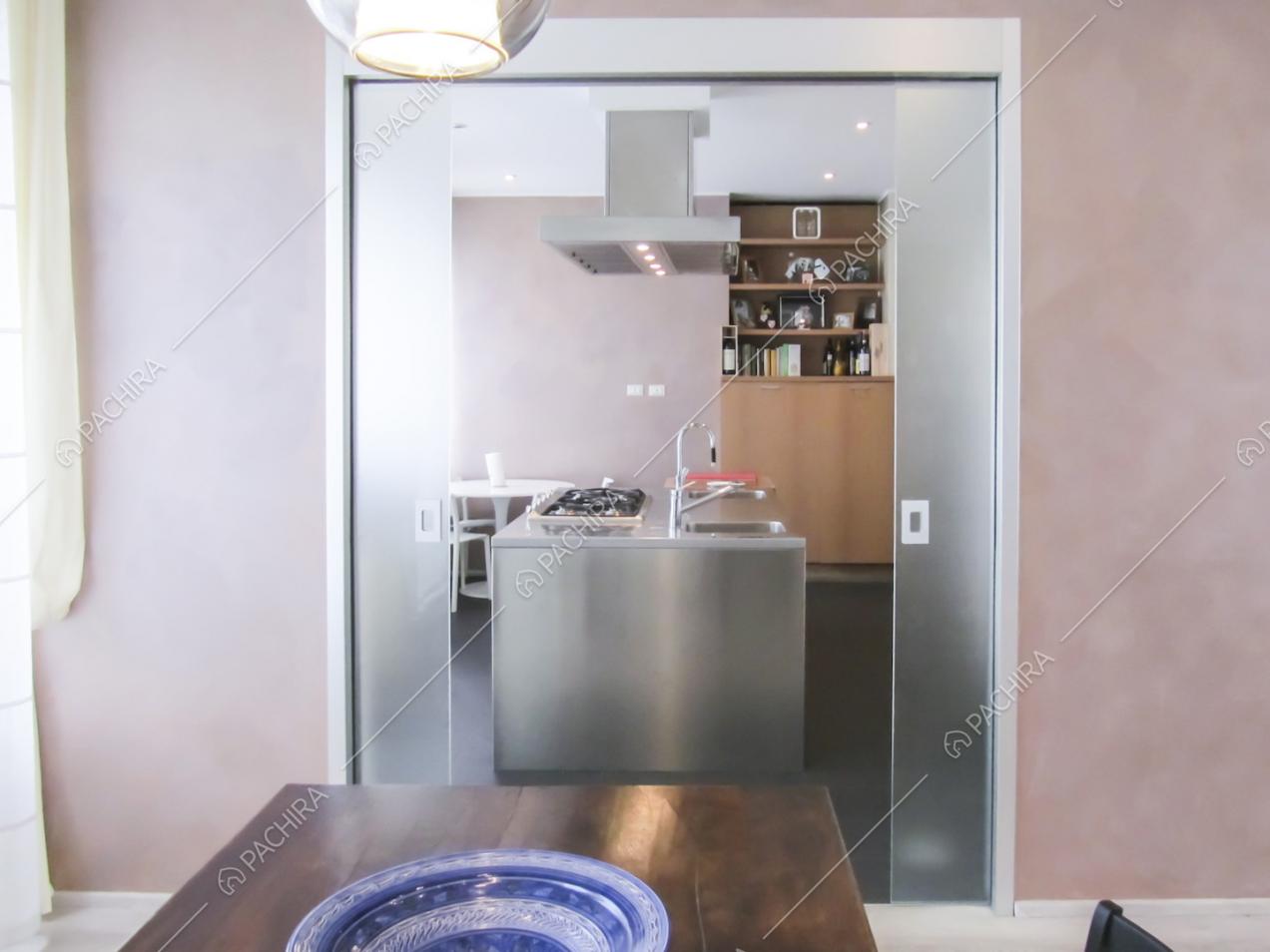 Porta doppia in vetro aperta, vista sull'isola della cucina dal tavolo da pranzo