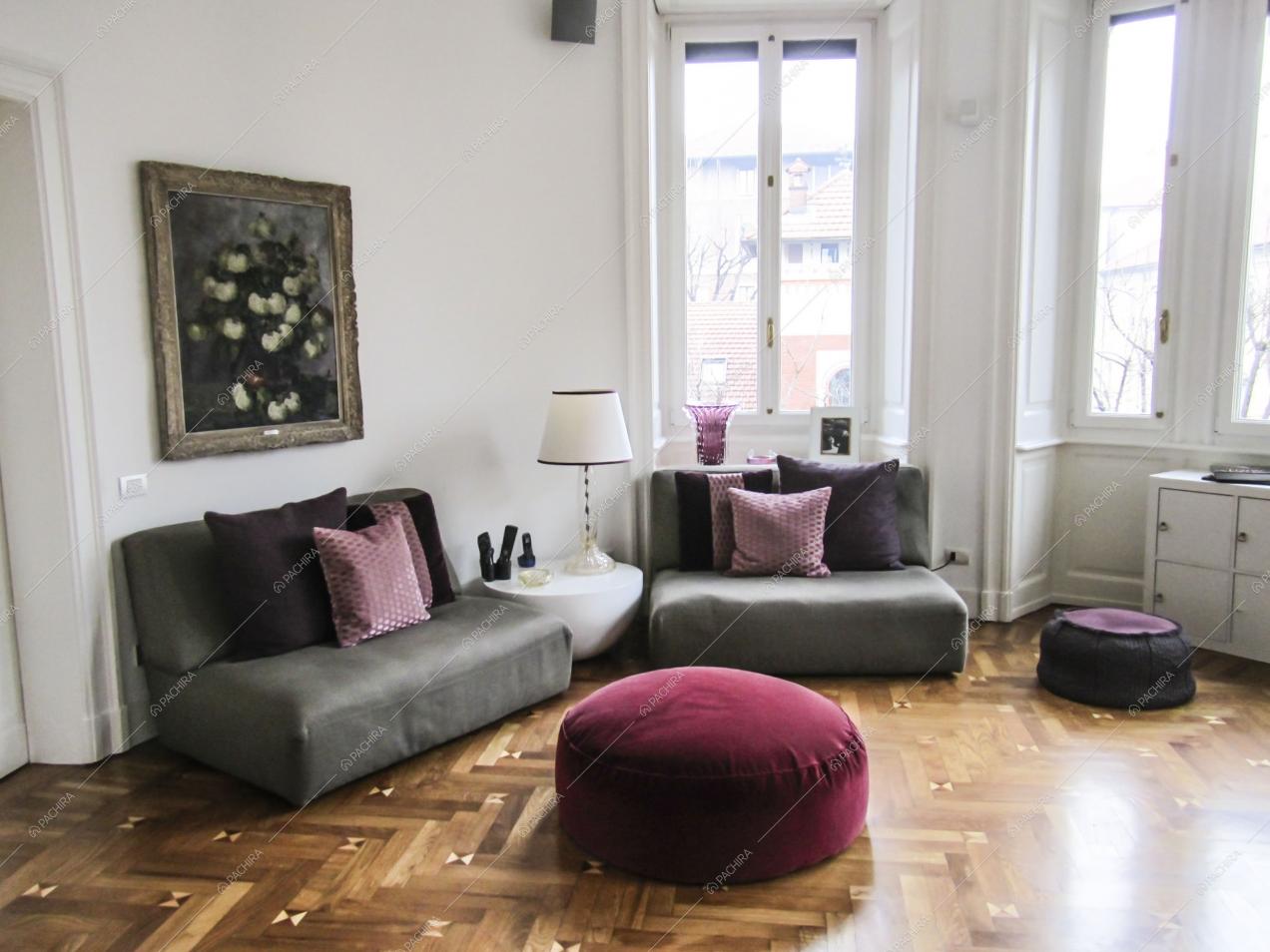 Luminoso soggiorno dalle pareti bianche con pavimento in parquet a spina di pesce, divani e pouf