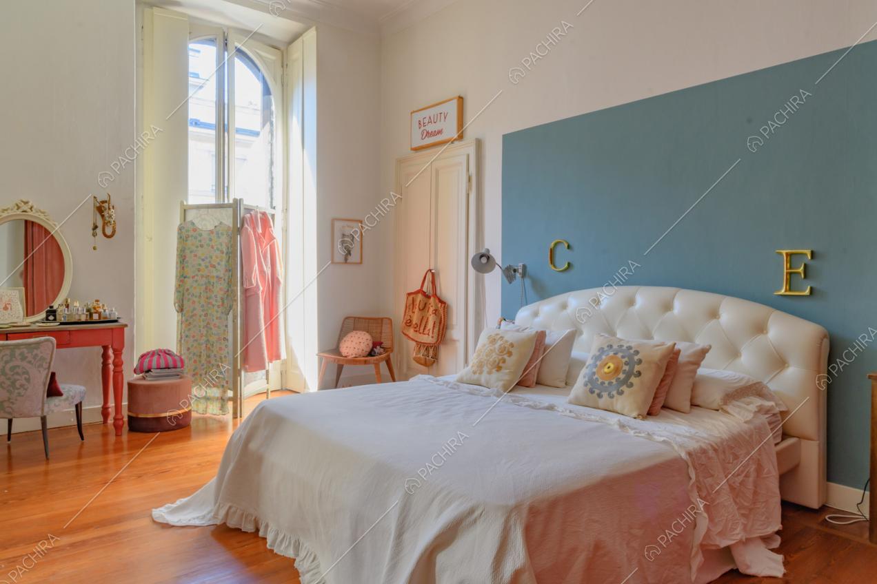 Camera da letto matrimoniale con testiera in pelle bianca, parete azzurra e pavimento in parquet